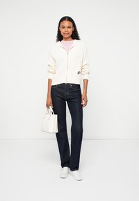 Kremowy dziergany kardigan na zamek z prążkowaną teksturą, połączony z ciemnoniebieskimi dżinsami. Akcesorium: mała biała torebka. Obuwie: białe sneakersy.