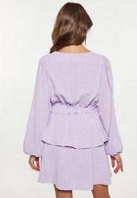 Rochie lavandă, ușoară, dintr-un material texturat, cu mâneci bufante lungi, talie peplum și fustă evazată. Vederea din spate arată detalii plisate.