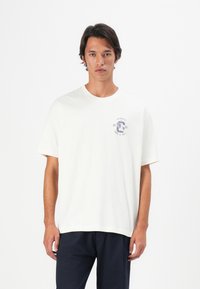 Junger Mann trägt ein weißes T-Shirt mit kurzen Ärmeln und einem kleinen Aufdruck "Los Angeles Track & Field 1981", kombiniert mit dunklen Hosen.