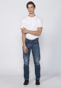 Una camiseta blanca de manga corta combinada con jeans azules desgastados. Los jeans tienen un corte ajustado, y el modelo lleva zapatillas negras.