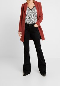 Abrigo color óxido, blusa negra con estampado de cebra y detalles de encaje, pantalones de mezclilla negros acampanados y zapatos negros de punta. Fondo liso.
