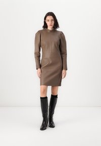 Rochie din piele maro cu mâneci lungi, având un guler înalt și umeri structurați, asortată cu cizme negre texturate până la genunchi.