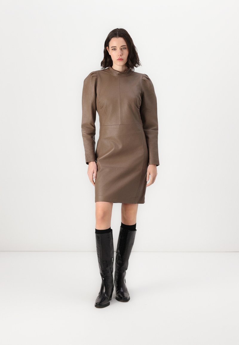 Rochie din piele maro cu mâneci lungi, având un guler înalt și umeri structurați, asortată cu cizme negre texturate până la genunchi.