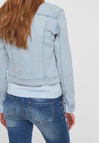 Vrouw met lang bruin haar, gekleed in een lichtblauwe spijkerjacket en blauwe spijkerbroek, staand tegen een effen witte achtergrond.