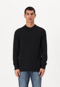 Svart strikkegenser med lange ermer, rund hals og ribbestrikkede mansjetter og kant. Brukt med denimjeans mot en ensfarget bakgrunn.