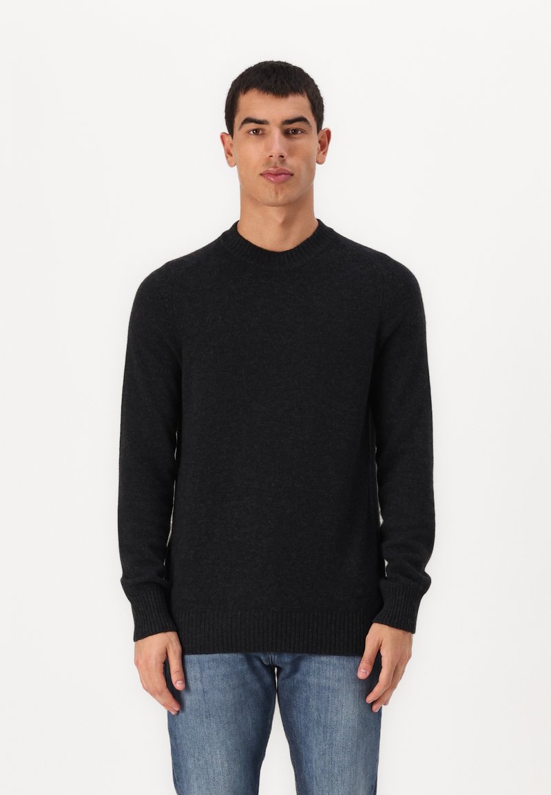 Svart strikkegenser med lange ermer, rund hals og ribbestrikkede mansjetter og kant. Brukt med denimjeans mot en ensfarget bakgrunn.