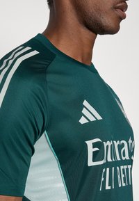 Mørkegrøn atletisk trøje med hvide accenter og branding. Glat tekstur med rund halsudskæring og skræddersyet pasform.