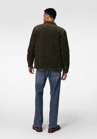 Mørkegrønn kordfløyelsjakke med krage, knappemanchetter og ryggpanel, kombinert med lyseblå jeans og brune sko.