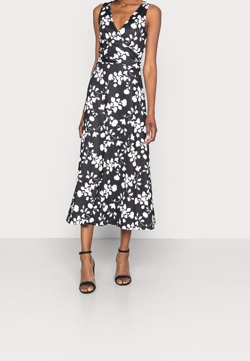 Svart blommig maxiklänning med vitt blommönster, V-ringning, ärmlös, flödande tyg och åtsittande midja. Bärs med svarta strappy klackar.