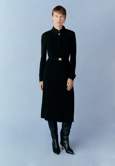 Black long-sleeve dress with a collared neckline and a belt at the waist. Material appears smooth; model wears knee-high black boots.