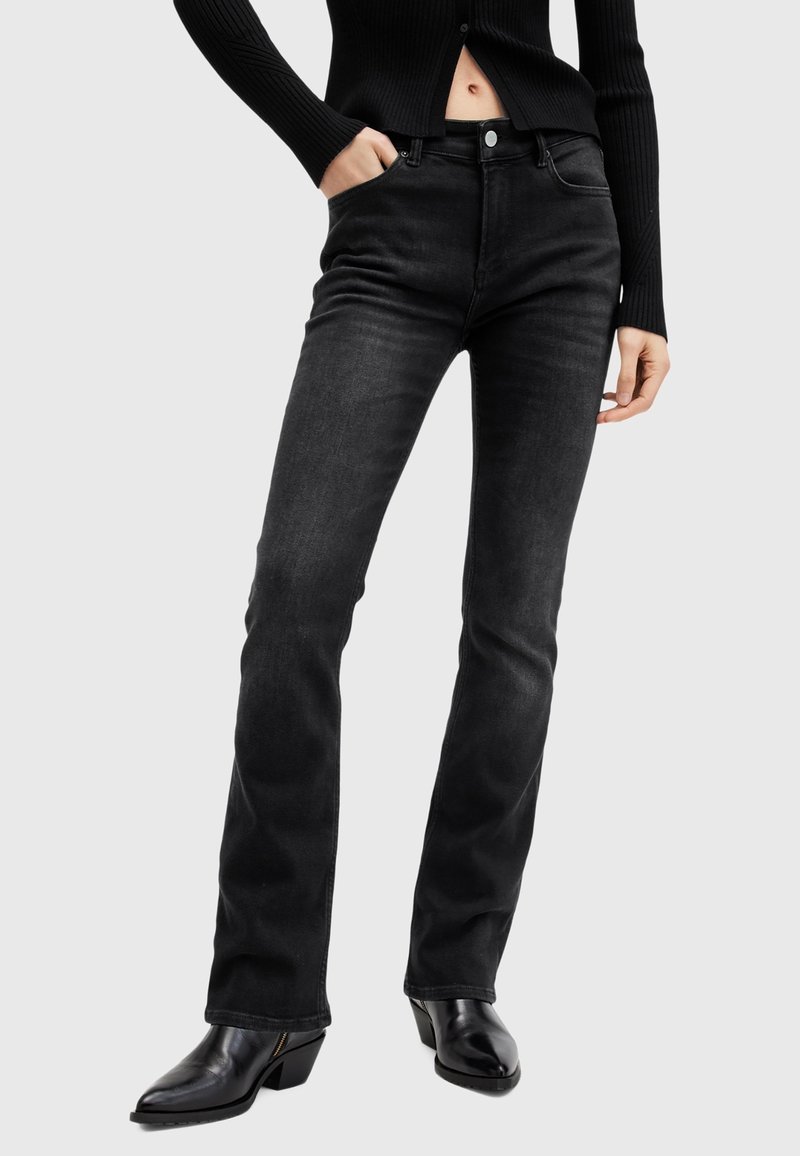 Personne portant un jean noir ajusté, des bottes noires à talons et un pull court noir côtelé, une main dans la poche.
