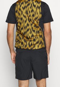 Schwarzes kurzärmeliges Hemd kombiniert mit einer mehrfarbigen, strukturierten Weste mit einem goldenen und schwarzen Zickzackmuster, getragen über dunklen Shorts.