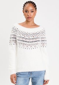Femme avec des cheveux tressés portant un pull blanc en laine avec des motifs géométriques noirs et bruns sur la poitrine et un jean bleu clair.