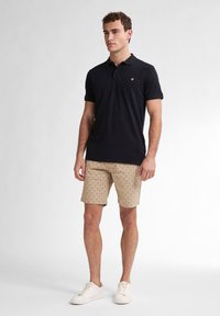 Polo nero, pantaloni corti beige con motivo e sneakers bianche. La maglietta ha un colletto e un piccolo logo sul petto.