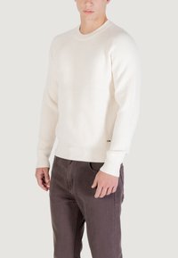 Maglione a coste di colore crema con scollo rotondo e maniche lunghe, abbinato a pantaloni marrone scuro. I dettagli significativi includono il tessuto testurizzato.