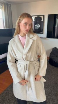 Ung kvinde med langt blondt hår iført en beige kåbe med bælte over en lyserød sweater, stående indendørs i en moderne stue.