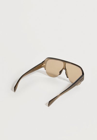 Καφέ αποχρωματισμένα ορθογώνια γυαλιά ηλίου με χοντρό, διαφανές σκελετό και λογότυπο Gucci στο πλαϊνό μέρος, εμφανιζόμενα από πίσω.