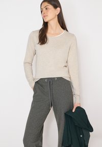 Femme debout en haut beige à manches longues et pantalon gris à cordon, tenant une veste pliée vert foncé dans une main.
