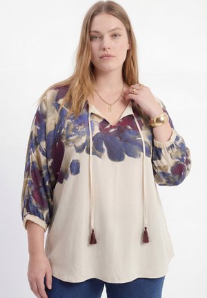 Vrouw met lang blond haar, gekleed in een crèmekleurige blouse met mouwen met een bloemenpatroon in blauw en paars, en gouden sieraden, staand tegen een witte achtergrond.