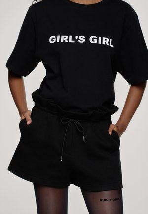 Personne portant un ensemble T-shirt noir et short avec le texte "GIRL'S GIRL" sur le haut et le même texte sur des collants noirs transparents.