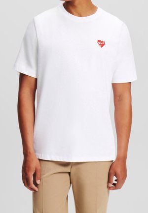 Weißes Baumwoll-T-Shirt mit kurzen Ärmeln, das ein kleines rotes Herzmotiv auf der Brust zeigt. Schlichte, gerade geschnittene Form mit Rundhalsausschnitt.