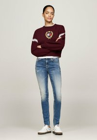 Burgundijas krāsas ikdienīgs svārku tops ar emblemu, baltās svītras uz piedurknēm, kombinēts ar ziliem, nolietotiem skinny džinsiem un baltiem sneakers ar brūnām zolitēm.