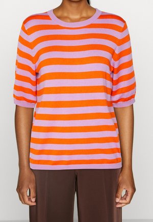 Person iført en kortærmet sweater med brede horisontale orange og lavendelfarvede striber, kombineret med mørkebrune bukser.