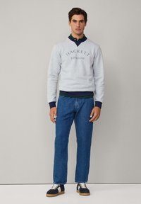 Sudadera gris con el logo "HACKETT LONDON", puños azul marino, superpuesta a un polo verde; combinada con vaqueros azules y zapatillas negras con suelas beige.