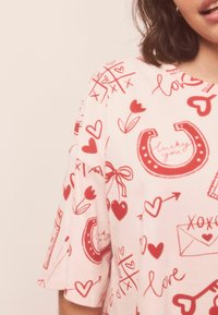 Rosa kortærmet T-shirt med røde mønstre, der forestiller hjerter, blomster, pile og en hestesko med teksten "heldige dig." Blød stoftekstur.