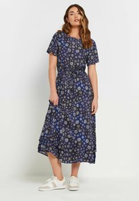 Marinblå blommig klänning med korta ärmar, har mönster av vita och blå blommor, smal midja och lager-på-lager nederdel. Kompletteras med vita sneakers.