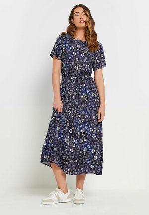 Vestido floral navy con mangas cortas, presenta patrones de flores blancas y azules, cintura ceñida y dobladillo en capas. Combinado con zapatillas blancas.