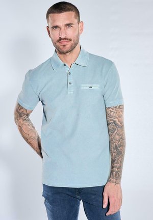 Mann mit kurzen Haaren und Bart, der ein hellblaues Polohemd und dunkle Jeans trägt, zeigt Tätowierungen auf beiden Unterarmen vor einfarbigem Hintergrund.