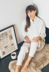 Gemütlicher cremefarbener Pullover mit geripptem Muster, weiße Hosen und barfuß. Eine gerahmte Zeitungsseite in Sepiatönen und eine weiße Tasse auf einer schwarzen Fläche.