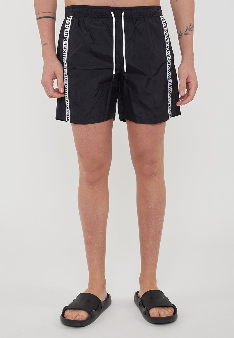 Pantaloni da bagno neri con strisce laterali bianche che riportano il testo del marchio. Tessuto leggero e ad asciugatura rapida con vita elastica e stringa.