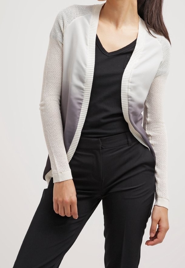 Femme portant un chemisier noir à col en V, un pantalon noir et un cardigan ouvert dégradé blanc à gris avec des manches en maille.