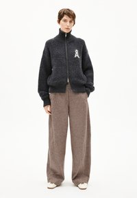 Sweat à fermeture éclair texturé foncé avec un col haut, arborant un logo blanc et des poignets côtelés, associé à un pantalon marron ample et des chaussures crème.