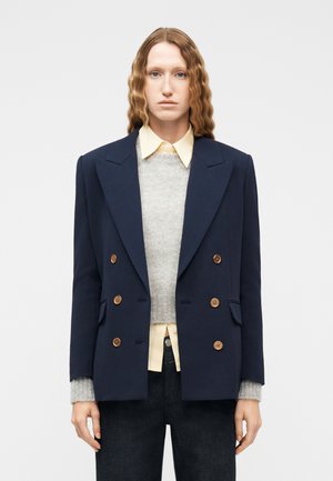 Vrouw met lang golvend haar, gekleed in een marineblauwe dubbelrijige blazer, een grijze trui, een lichtgele overhemd met kraag en een donkere broek, staand tegen een witte achtergrond.