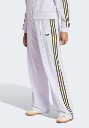 Pantaloni della tuta bianchi con tre strisce marroni ai lati e logo Adidas, indossati con giacca coordinata e sneakers beige.