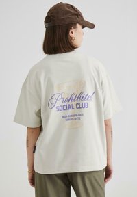 Light grey oversized t-shirt featuring purple and gold graphic text on the back, with short sleeves and relaxed fit fabric.