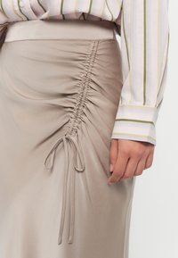 Beige Satinrock mit seitlichem Raffung und einer Bindeschleife. Kombiniert mit einem rosa-weißen Streifenhemd mit grünen Akzenten an den Manschetten.