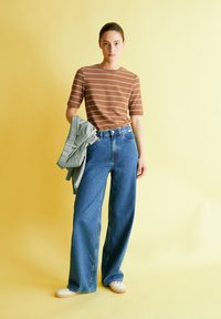 Bruine gestreepte kortleding shirt gecombineerd met high-waisted, wijde blauwe denim jeans. Model houdt een lichtgrijze trui vast. Crème-kleurige gevlochten schoenen.