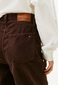 I pantaloni in corduroy marrone presentano un'etichetta in pelle con la scritta "TOMMY JEANS", tasche posteriori e accenti di colore sottili nella cucitura e nel logo.