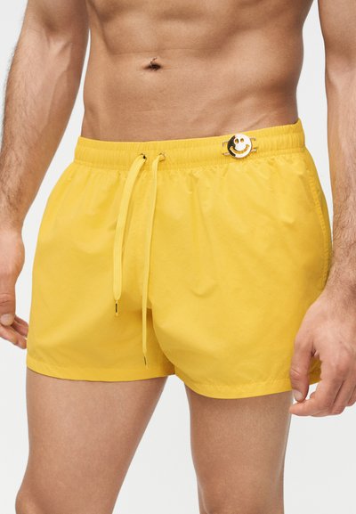 Άνδρας που φοράει κίτρινα μαγιό με κορδόνι και μικρό μεταλλικό καρφίτσα με χαμογελαστό προσωπάκι στη ζώνη, δείχνοντας τον τονωμένο κορμό και τα μπράτσα του.