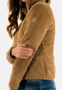 Giacca in pile color tan con un motivo testurizzato a pois, colletto alto, chiusura con zip e un piccolo patch con logo sulla spalla.