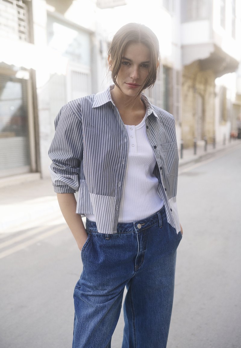 Mlada žena stoji na mirnoj ulici, odjevena u skraćenu prugastu majicu preko bijelog topa i labavim plavim trapericama, s rukama u džepovima.