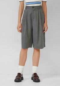 Persona che indossa pantaloni corti grigio a righe verticali fino al ginocchio, calze bianche e mocassini in pelle marrone scuro, in piedi contro uno sfondo neutro.