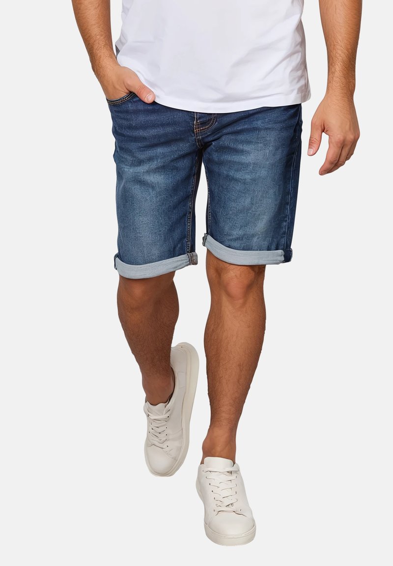 Mann trägt hochgekrempelte blaue Jeansshorts, weiße Sneakers und ein weißes T-Shirt, geht mit einer Hand in der Tasche.