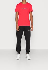 Camiseta de algodón roja con el logo "HILFIGER", combinada con pantalones de chándal negros que tienen un parche con logo, y completada con zapatillas blancas.