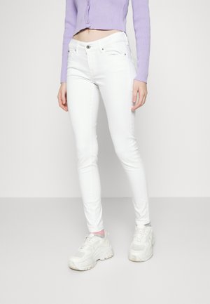 Vrouw draagt witte slim-fit jeans, witte chunky sneakers en een cropped lavendelkleuren cardigan, staand tegen een eenvoudige achtergrond.