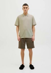 Koszulka polo z beżowego tkanego materiału z kołnierzykiem i krótkimi rękawami, zestawiona z brązowymi szortami ze spodenkami. Całość uzupełniają czarne buty.